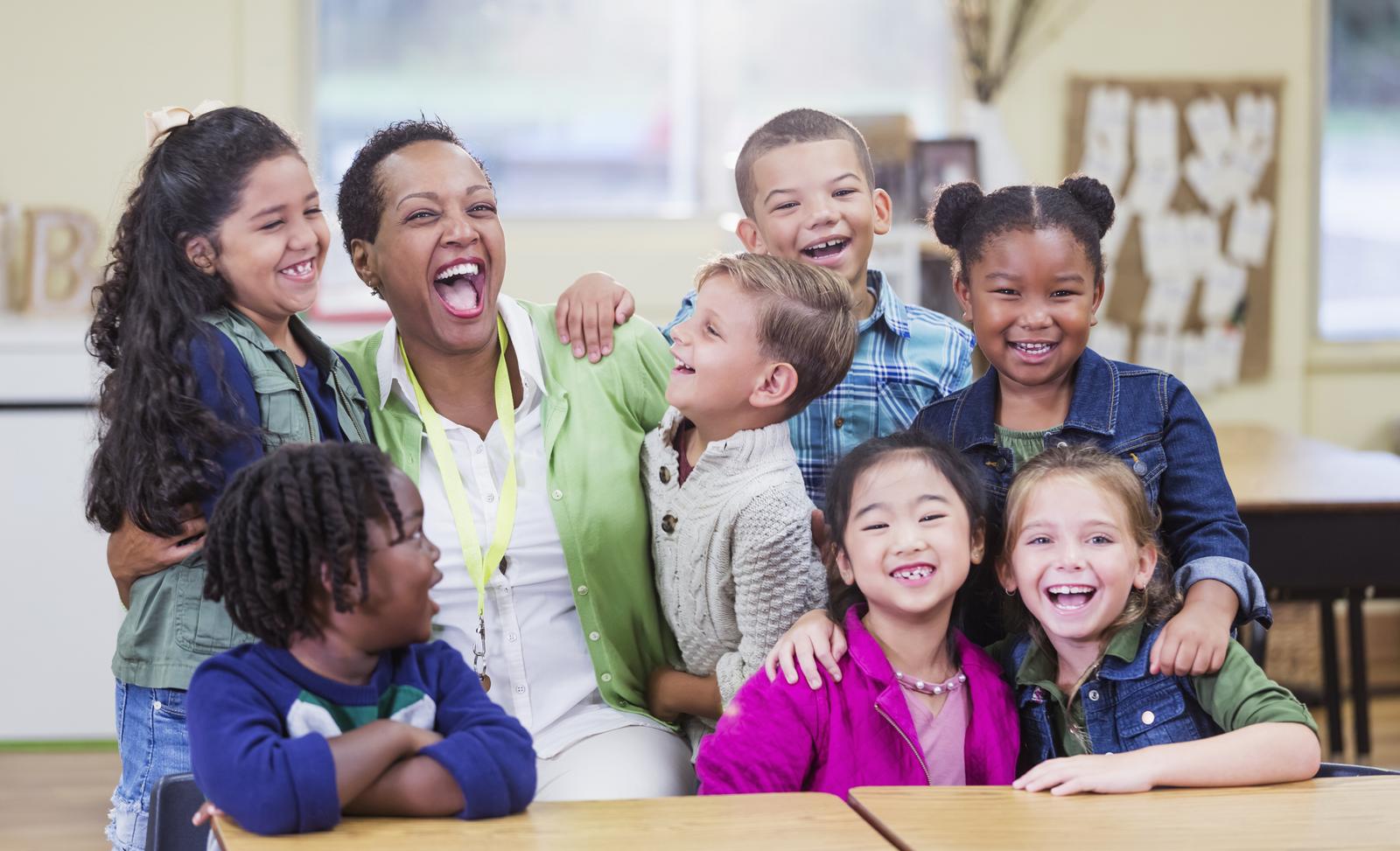 A teacher laughing with a group of elementary school students in a bright classroom